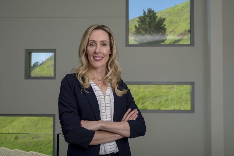 Blonde woman posing with arms crossed in front of a wall of windows.