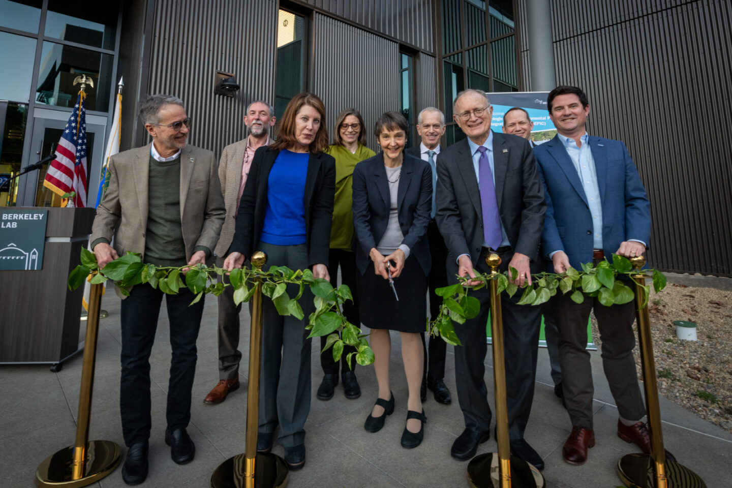 People gathered outside of a gray building, grouped behind a garland made of plants just after the central figure has made the ceremonial cut.