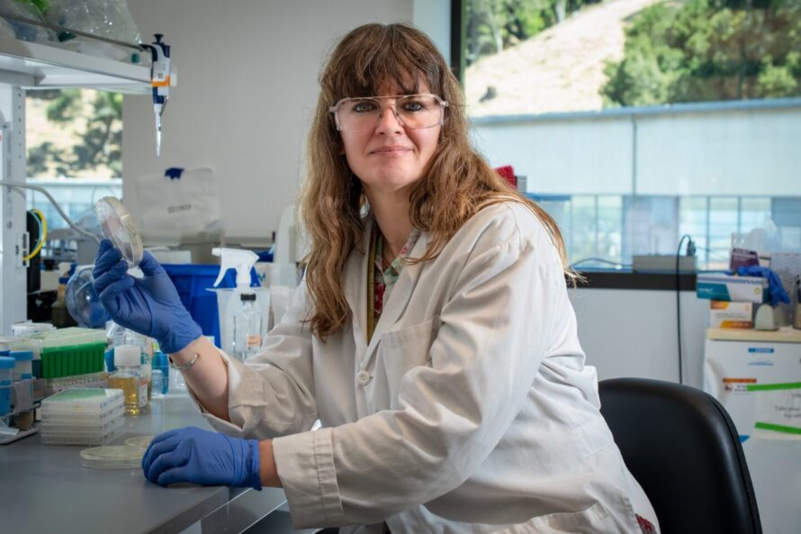 A scientist in a lab coat and safety goggles holds a petri dish at a laboratory bench.
