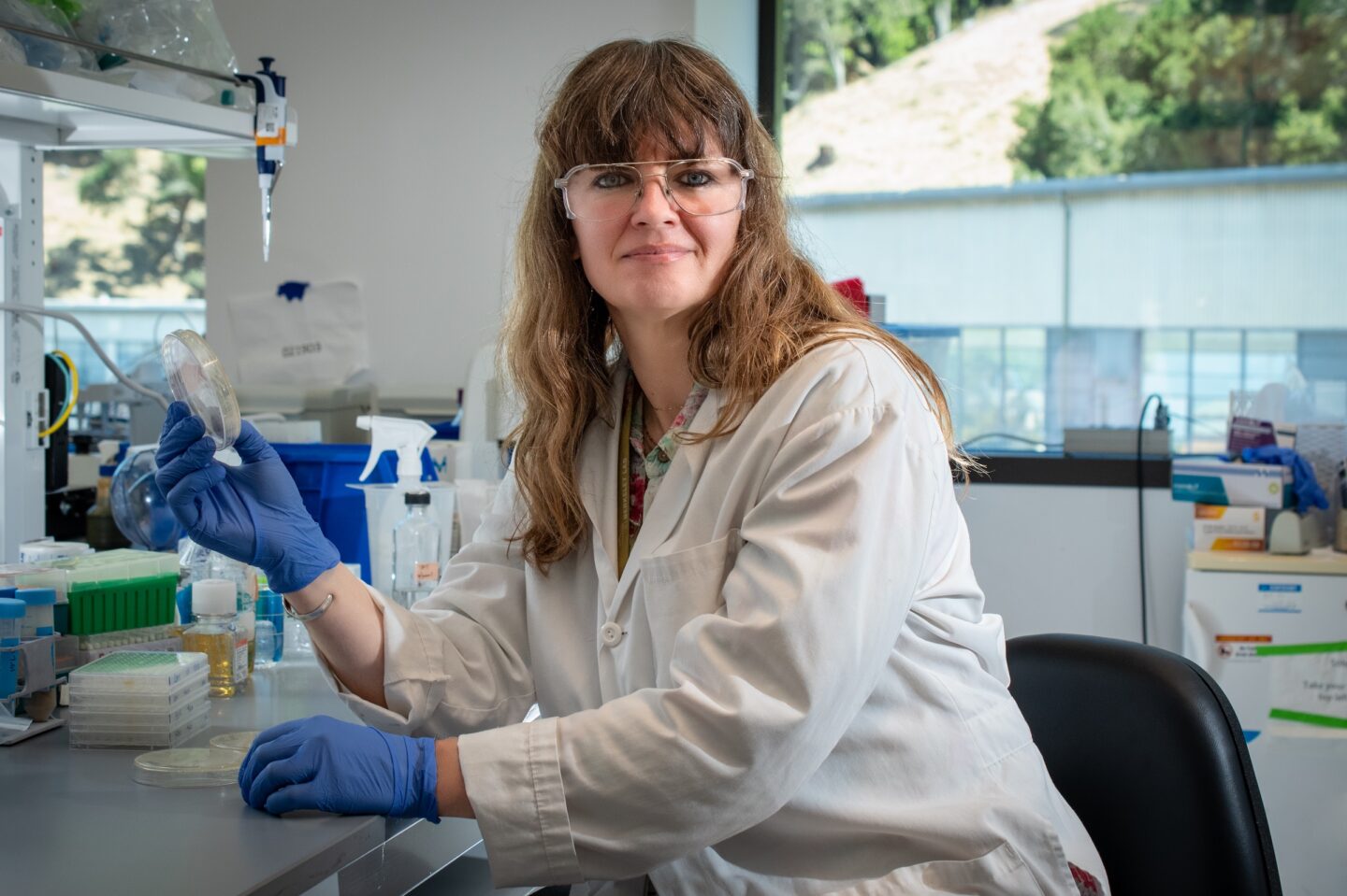 A scientist in a lab coat and safety goggles holds a petri dish at a laboratory bench.