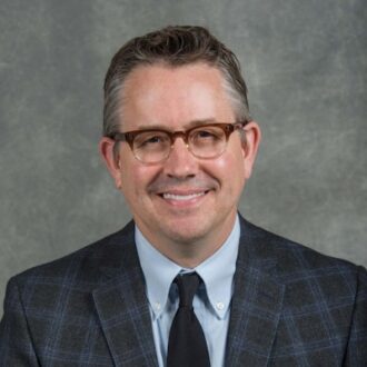 Man wearing a suit and tie, with dark glasses frames and dark hair.