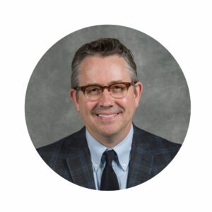 Man wearing a suit and tie with short dark hair and glasses.