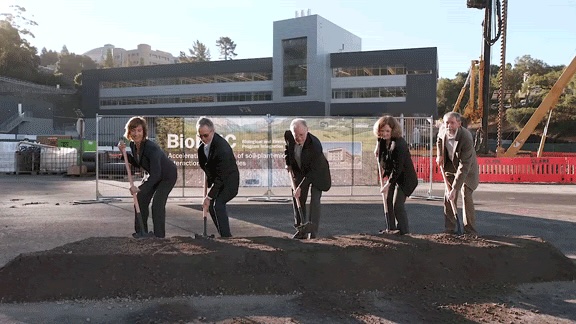 Five people use shovels in front of new building.